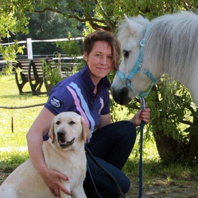 Frau kniet neben einem Hund und einem Pony auf einer Wiese.