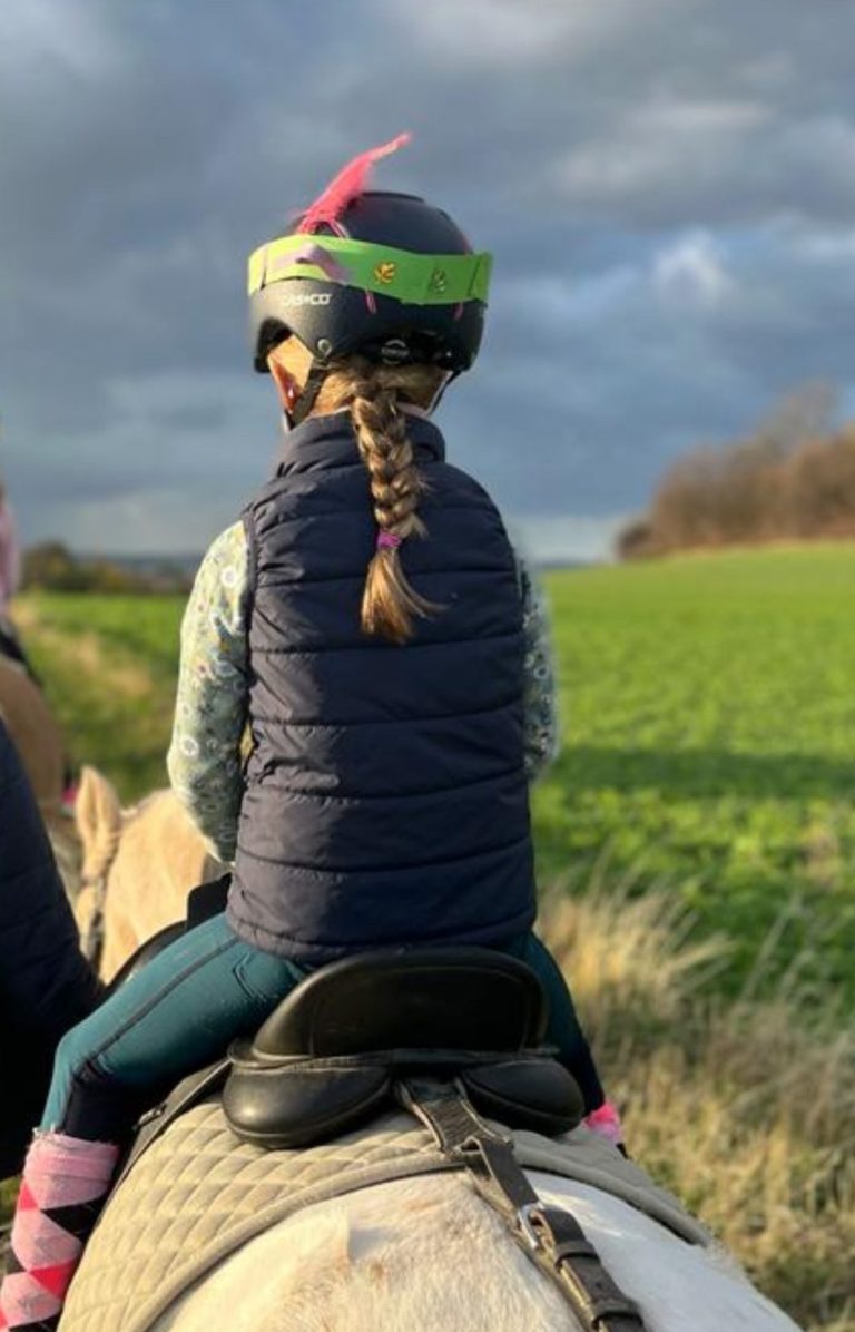 Mädchen mit einem bunten Helm reitet auf einem Pony, während Erwachsene und ein Kind begleiten.